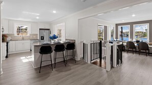 A modern kitchen with white cabinetry, a central island with bar stools, and a view of the outdoors through large windows. - Hillstone Ridge (Mooresville)