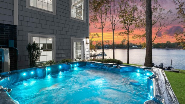 A hot tub with a view of a lake and sunset, a house with large windows, and a dock in the background. - Hillstone Ridge (Mooresville)
