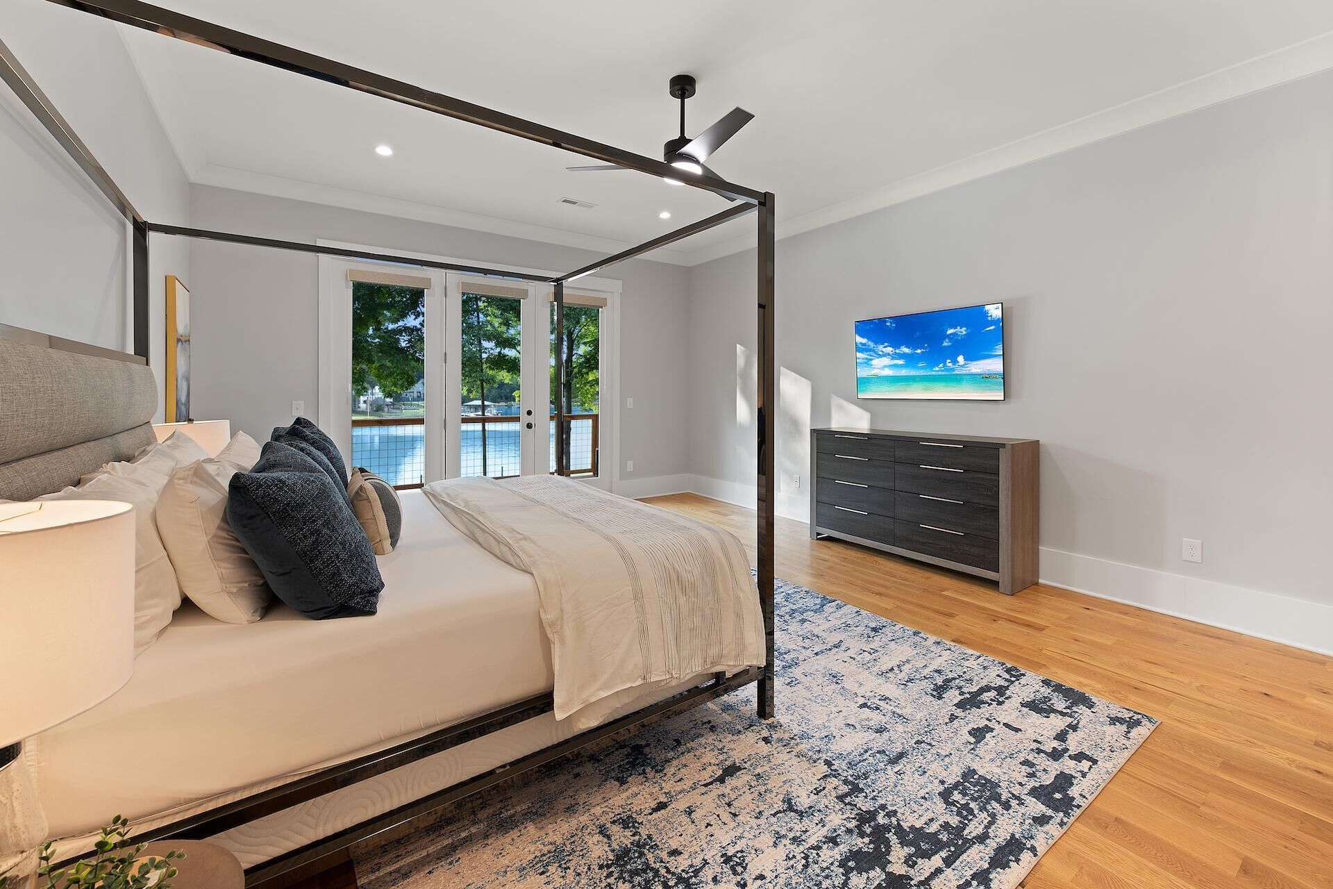 A bedroom with a four-poster bed, a nightstand, a dresser, and a large window with a view of a body of water.