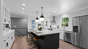 Cozinha moderna com ilha central, eletrodomésticos de aço inoxidável, iluminação pendente e backsplash em padrão herringbone.