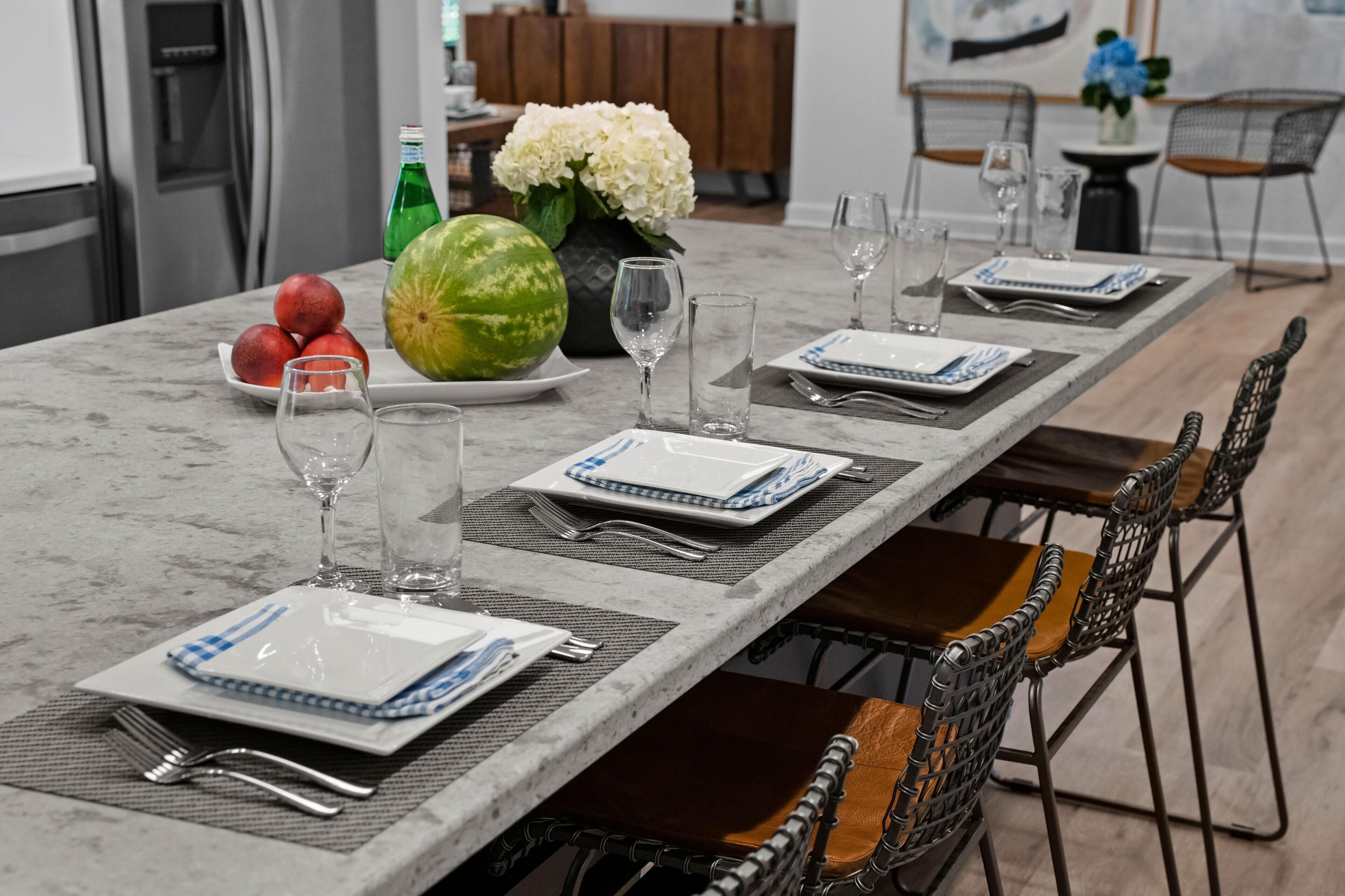 A dining table set with plates, glasses, and cutlery, with a watermelon and apples on the side.