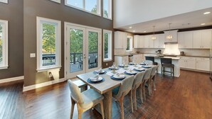 Una cocina amplia con armarios blancos, un comedor con una mesa de madera grande dispuesta para seis personas, y ventanas grandes que ofrecen vistas a árboles.