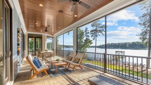 A spacious covered deck with wooden flooring, a ceiling fan, and large windows offering a view of a lake and trees. - Mallard Vestige (Denver)
