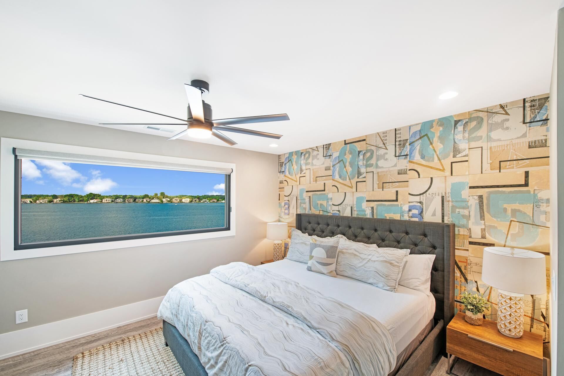 A bedroom with a large window, a ceiling fan, a bed with a tufted headboard, and a wall with abstract art.