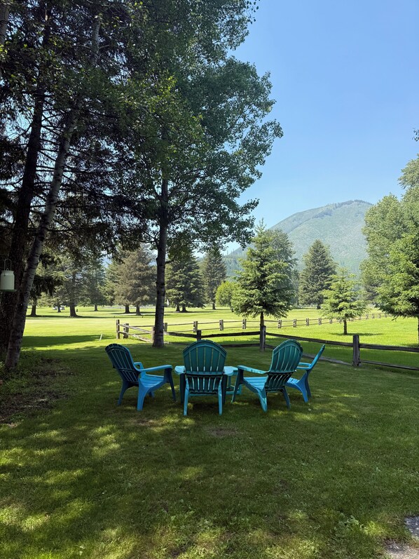 A grassy area with four blue Adirondack chairs arranged in a circle, surrounded by trees and a wooden fence. - West Glacier Hot Tub! On Golf Course! Private Home (Flathead County)