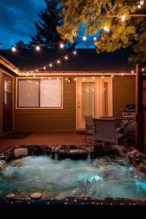 A hot tub with a wooden deck, string lights, and a door leading to a covered patio area. - West Glacier Hot Tub! On Golf Course! Private Home (Flathead County)