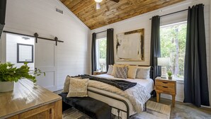 A bedroom with a bed, bedside table, bench, and a window with curtains. - Summerwoods: Sleeps 8 | Fire Pit | Hot Tub | EV (Broken Bow)