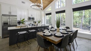 A modern kitchen with white cabinetry, a central island with seating, and large windows offering a view of the forest. - Summerwoods: Sleeps 8 | Fire Pit | Hot Tub | EV (Broken Bow)