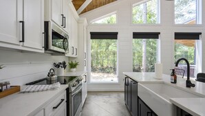 A modern kitchen with white cabinetry, stainless steel appliances, and large windows offering views of the outdoors. - Summerwoods: Sleeps 8 | Fire Pit | Hot Tub | EV (Broken Bow)