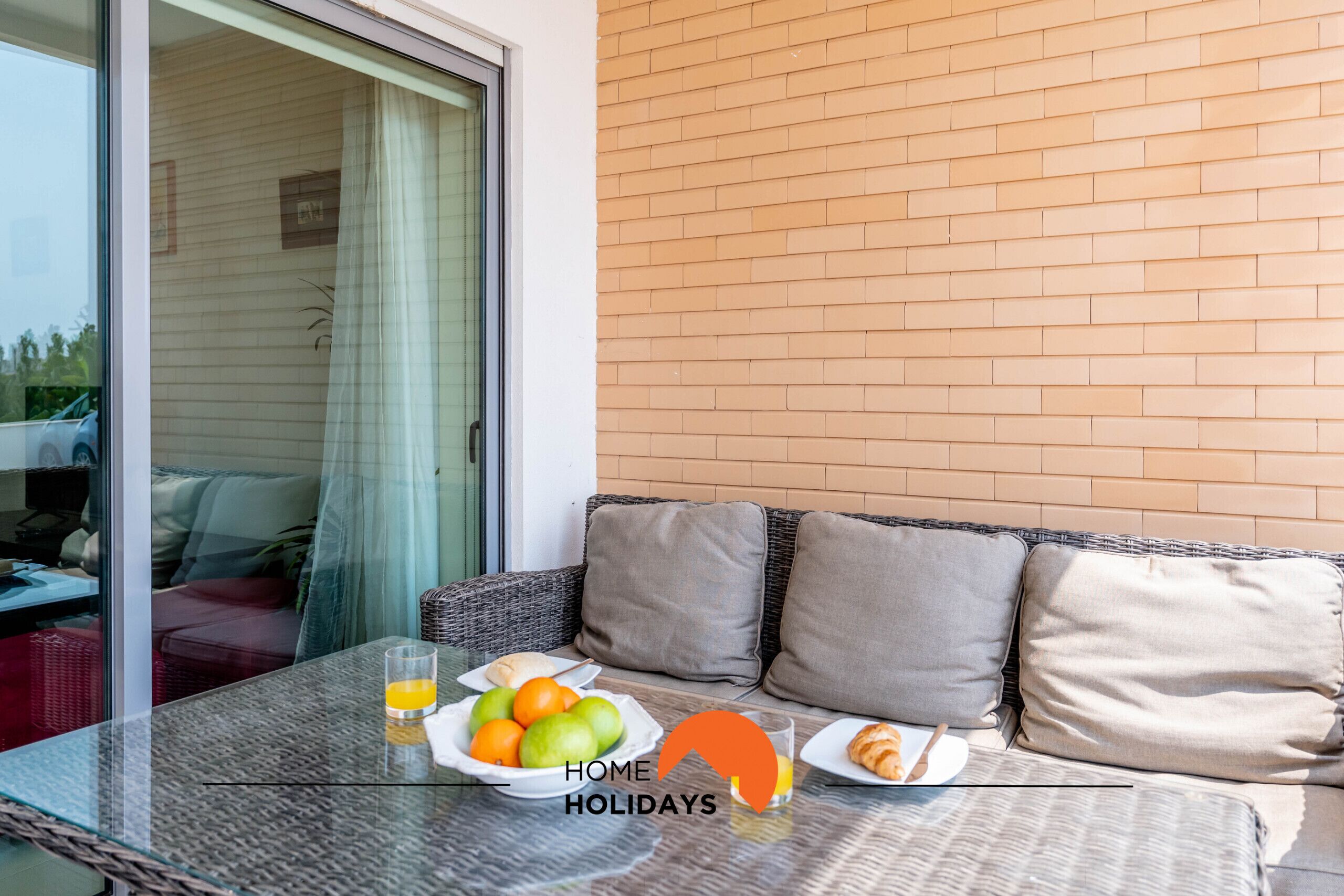 Une table avec un plateau en verre, un panier de fruits, un verre de jus d’orange et une assiette de croissants.