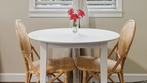 A round white table with wicker chairs, a vase with red flowers, and a window with white blinds and beige curtains. - Main Street Charmer 3 Bedrooms (New Iberia)