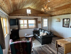 Ein gemütliches Blockhaushaus-Interieur mit Holzbalkendecke, einem schwarzen Ledersofa, einem gemusterten Sessel, einem kleinen runden Tisch und einem Holznachttisch.