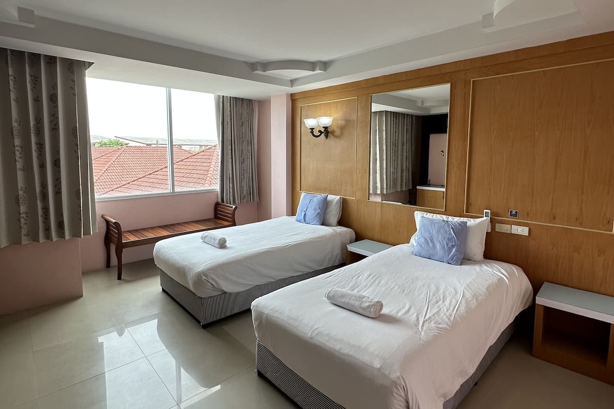 A hotel room with two beds, a bench, a large window, and a wooden wardrobe.