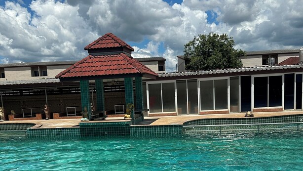 Une piscine à l’eau claire, un kiosque et un bâtiment avec un toit rouge en arrière-plan.