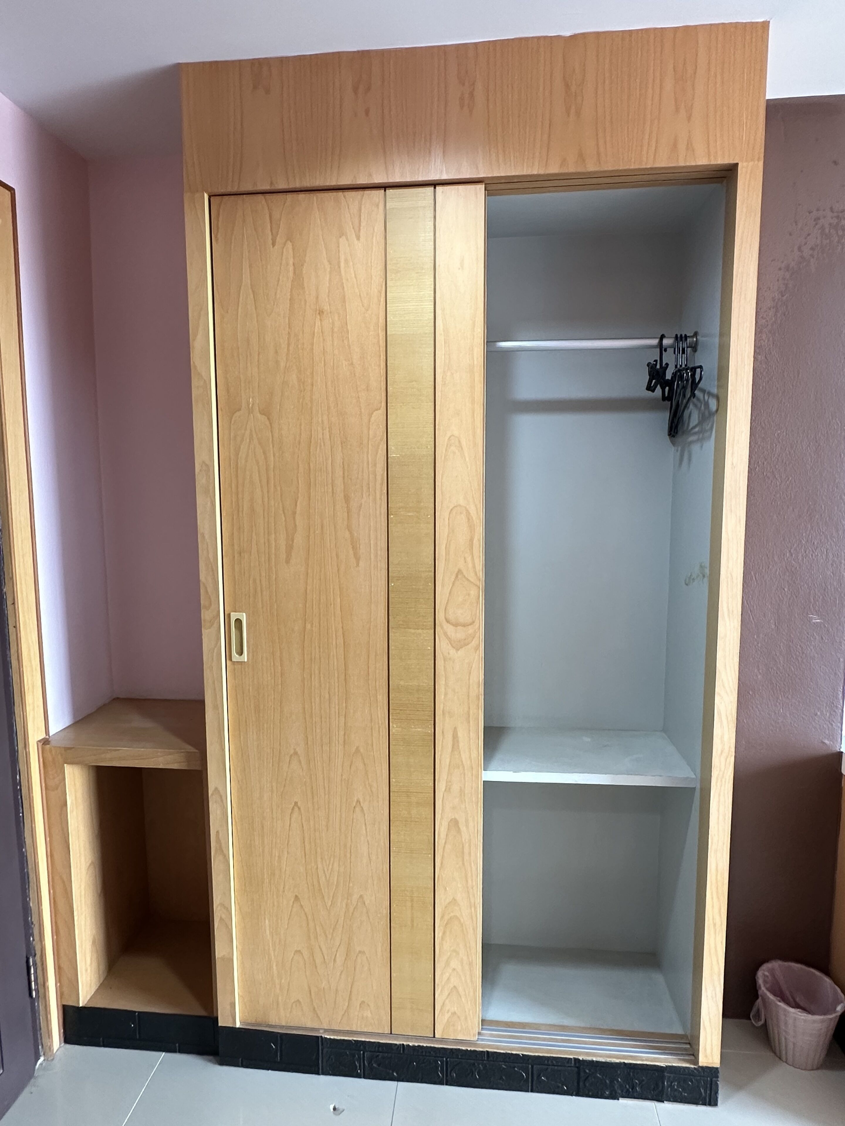 A wooden wardrobe with a sliding door and a shelf, located in a room with a tiled floor and a pink wall.