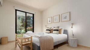 Un dormitorio con cama, una silla, mesita de noche con lámpara y ventana con vistas a follaje.
