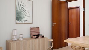 Una habitación con puerta de madera, una mesita, un mueble con adornos y una planta enmarcada en la pared.