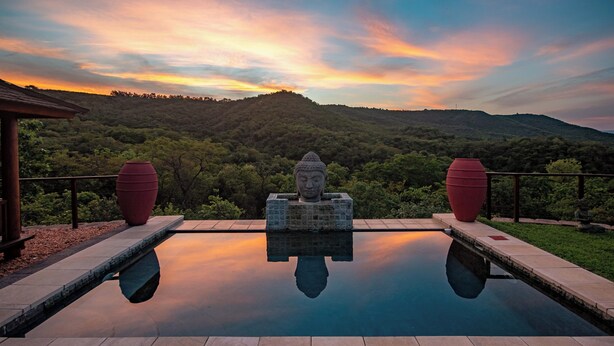 Un área de piscina tranquila con una estatua de Buda en el centro, rodeada de exuberante vegetación y un pintoresco paisaje de montañas.