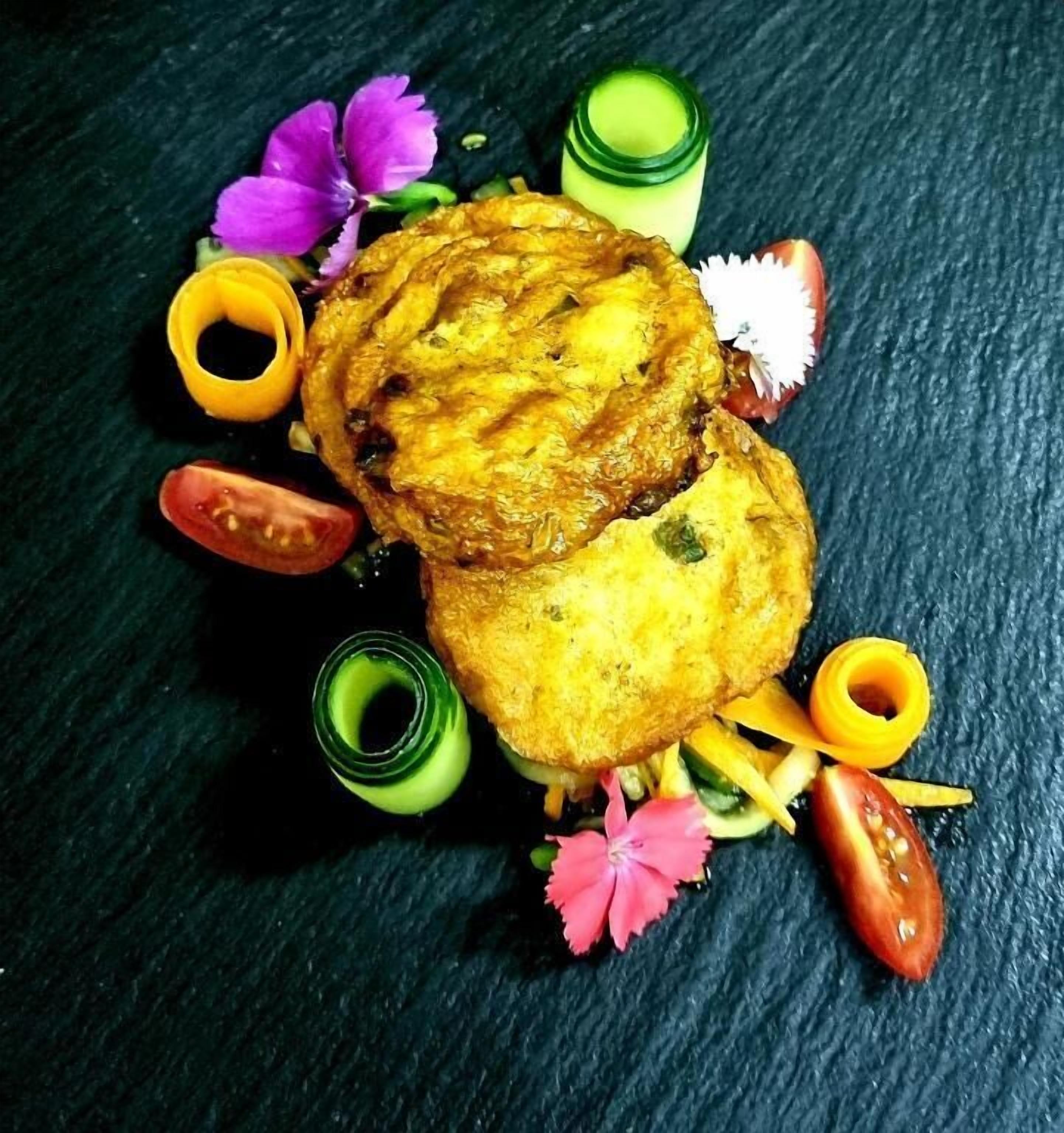 A dish with a golden-brown crust, surrounded by colorful garnishes and a dark textured background.