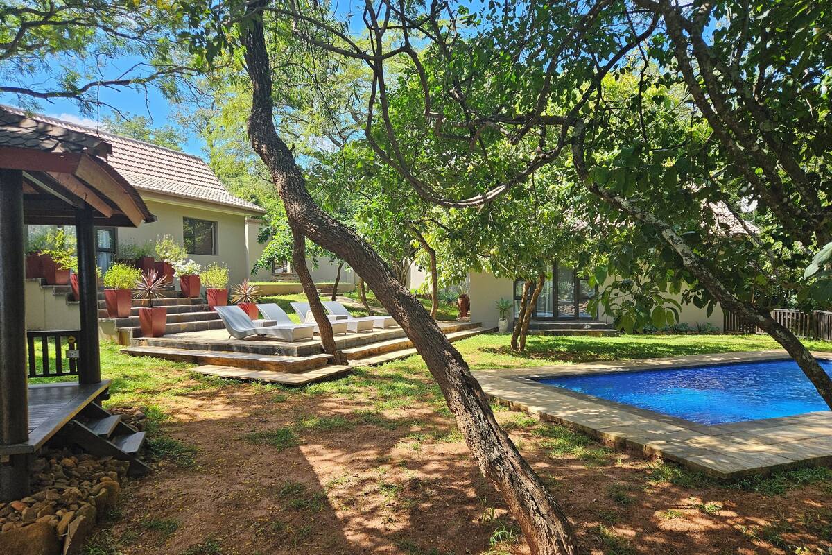 Ein Garten mit Swimmingpool, einer überdachten Terrasse mit Liegestühlen und einem Haus mit Ziegeldach.
