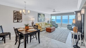 A spacious living room with a dining area, large windows offering a view of the ocean, and a ceiling fan. - Beach View 1 Bedroom 902 (Sarasota)