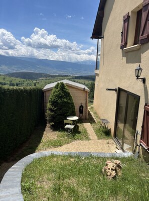 A small wooden shed with a table and chair in a garden area, a glass door, and a view of a mountainous landscape.