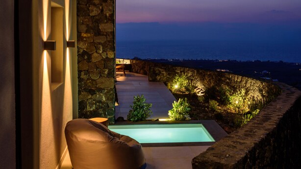 A modern outdoor seating area with a stone wall, a pool, and a view of the sea at dusk. - RyoLithos Suites (Santorini)