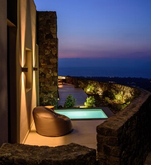 A modern outdoor seating area with a stone wall, a pool, and a view of the sea at dusk. - RyoLithos Suites (Santorini)