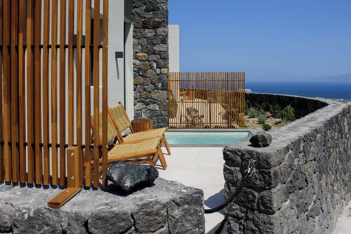 Zona de piscina con sillas de madera y un muro de piedra, con vista al mar.
