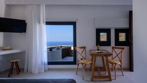 A room with a large window overlooking the sea, a table with chairs, a flat-screen TV, and a bed. - RyoLithos Suites (Santorini)
