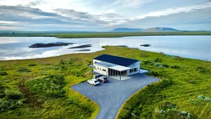 A modern house with a large garage and a driveway, surrounded by lush green fields and a lake.