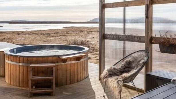 A wooden deck with a hot tub and a chair, overlooking a beach and water body.