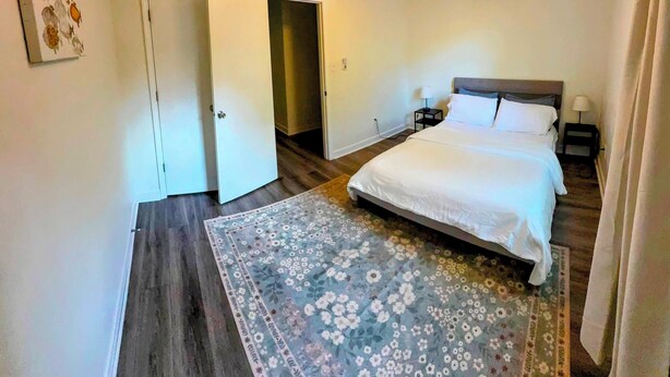 A bedroom with a bed, a ceiling fan, a nightstand, a wall-mounted air conditioner, and a door leading to another room.