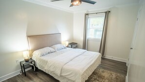 Een slaapkamer met een bed, nachtkastjes, een plafondventilator, een raam met gordijnen en een houten vloer.