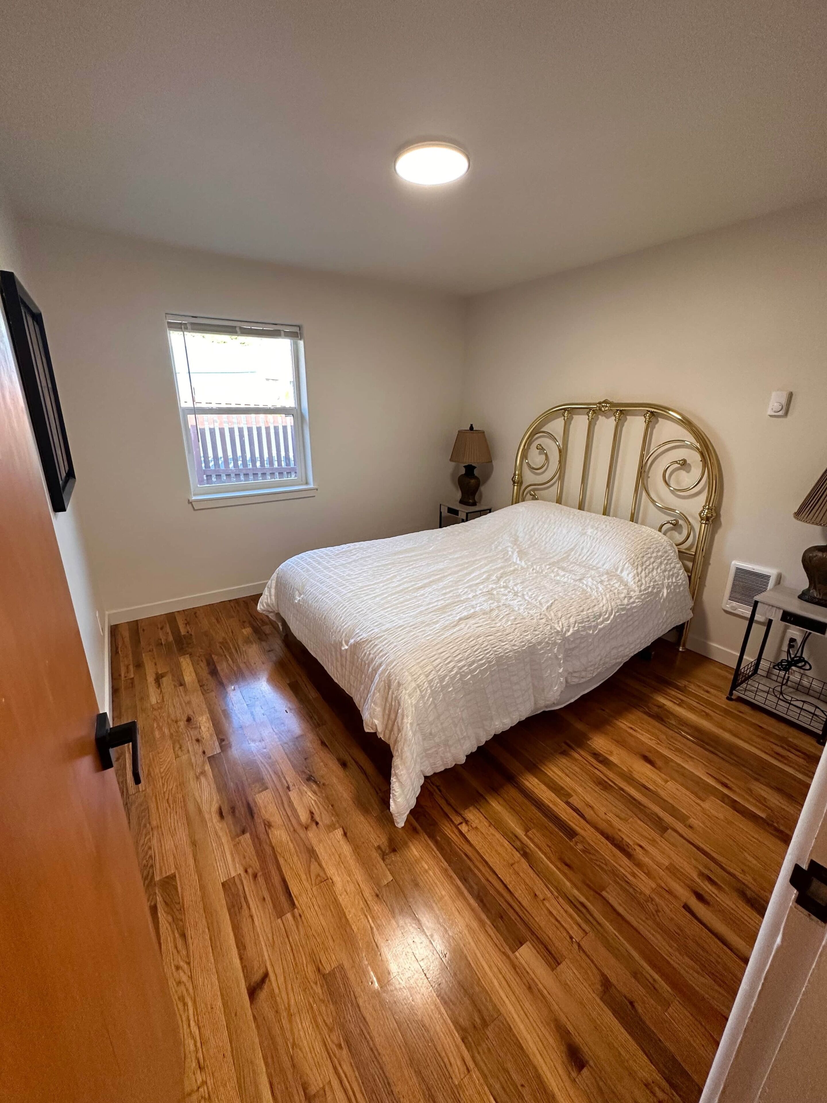 A bedroom with a wooden floor, a bed with a white bedspread, a window with a view, and a ceiling light.