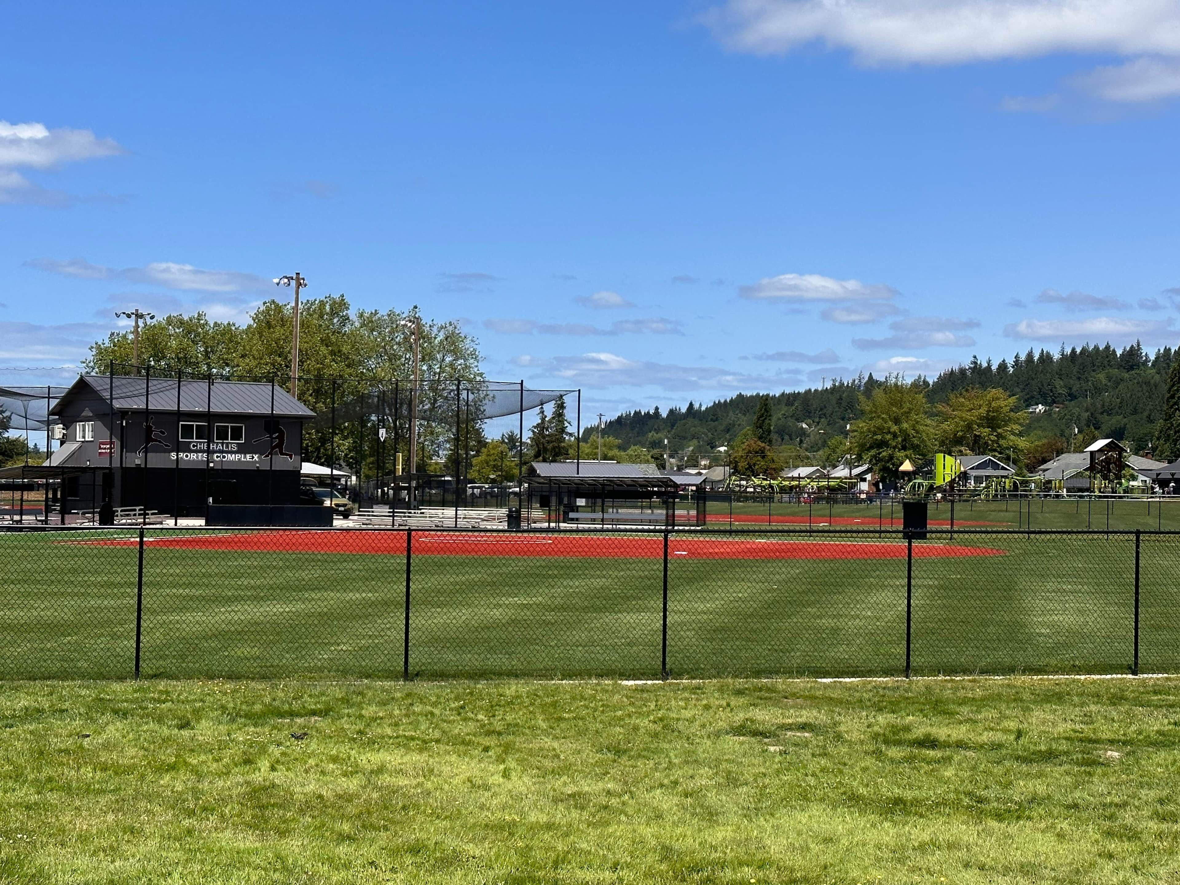 A sports complex with a fenced field, a building labeled 'CHE HALIS SPORTS COMPLEX,' and a forested hill in the background.