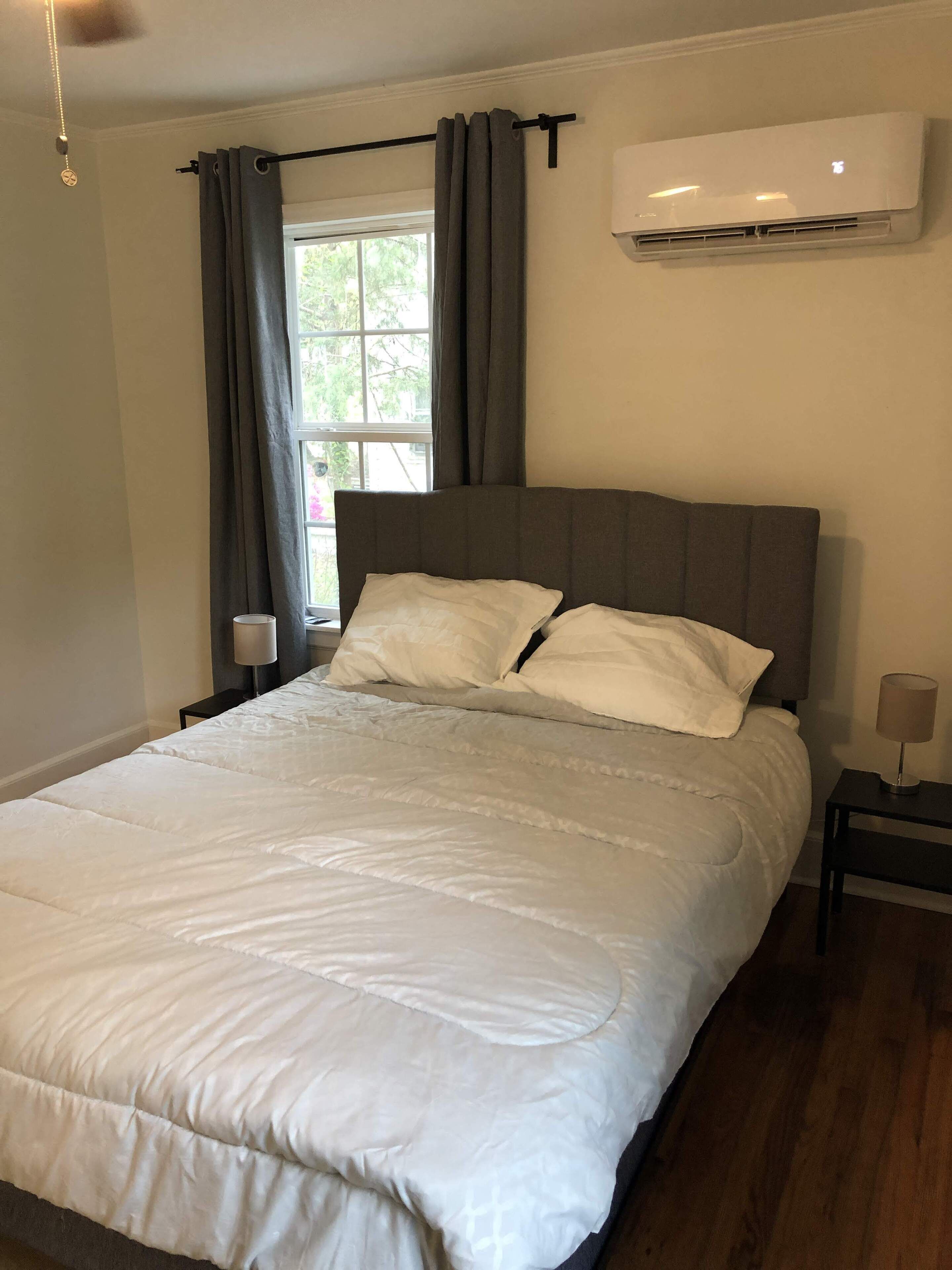 Un dormitorio con cama, ventana con cortinas, aire acondicionado y mesita de noche con lámpara.