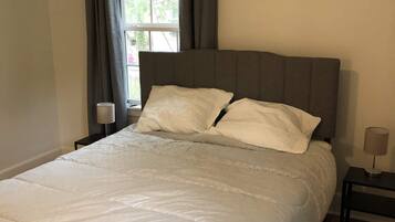 Un dormitorio con cama, ventana con cortinas, aire acondicionado y mesita de noche con lámpara.