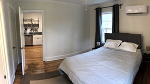 Ein Schlafzimmer mit Bett, Nachttisch, Lampe, einem Fenster mit Vorhängen und einer Klimaanlage.