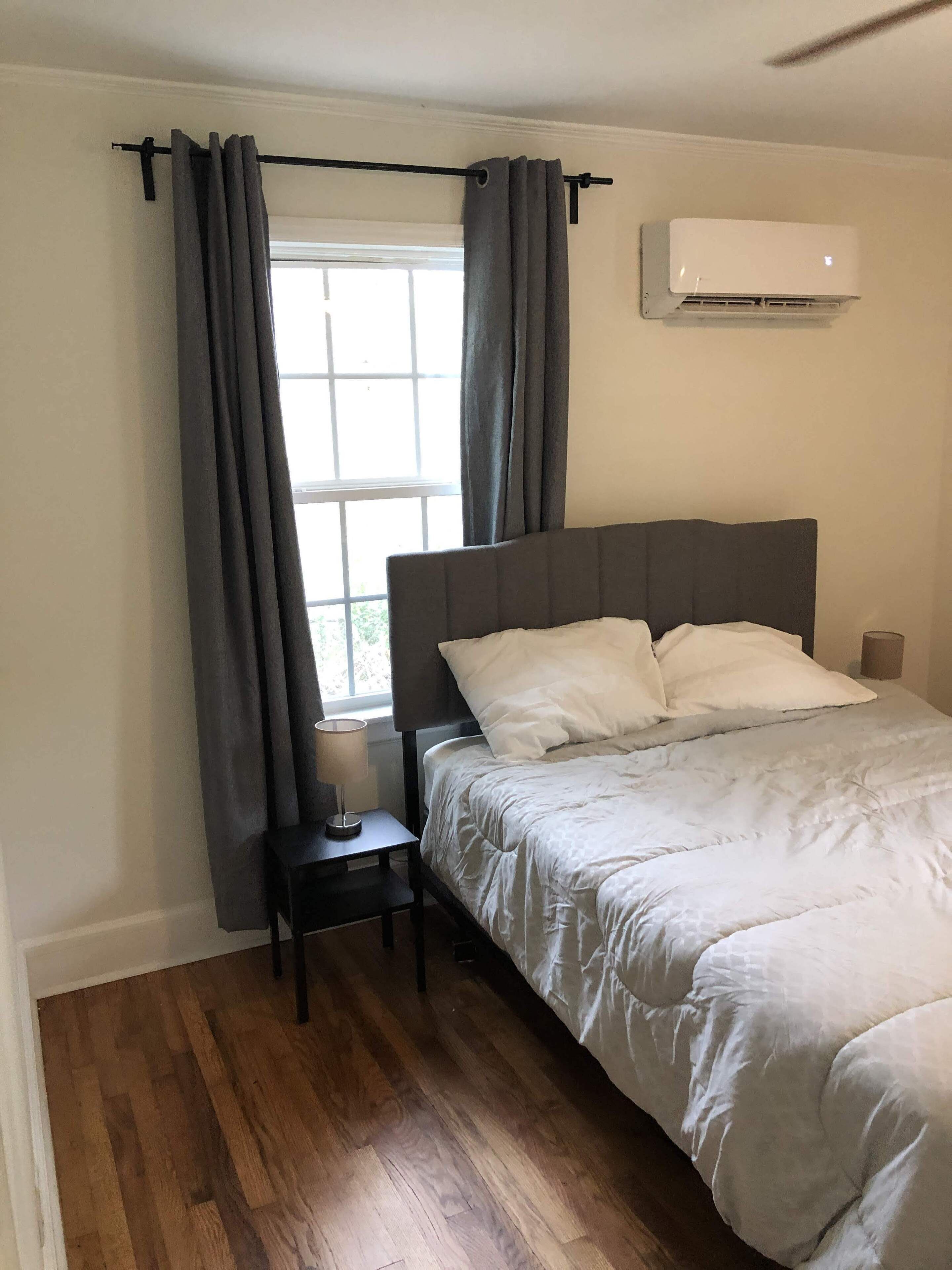A bedroom with a bed, a nightstand, a window with curtains, and an air conditioning unit.