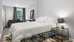 Ein modernes Schlafzimmer mit einem großen Bett, einem Nachttisch mit Lampe, einer Stehlampe und Blick auf Grünflächen.