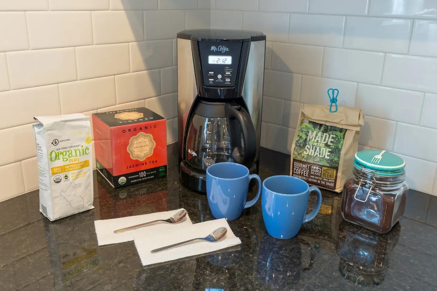 Eine Küchenarbeitsplatte mit einer Kaffeemaschine, einer Schachtel Bio-Tee, zwei blauen Tassen, einem Löffel, einer Packung schattengewachsenen Kaffees und einem Kaffeiglas.