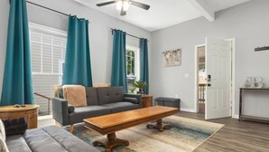 Ein geräumiges Wohnzimmer mit grauem Sofa, einem Holztisch, blaustichigen Vorhängen und einem Deckenventilator.