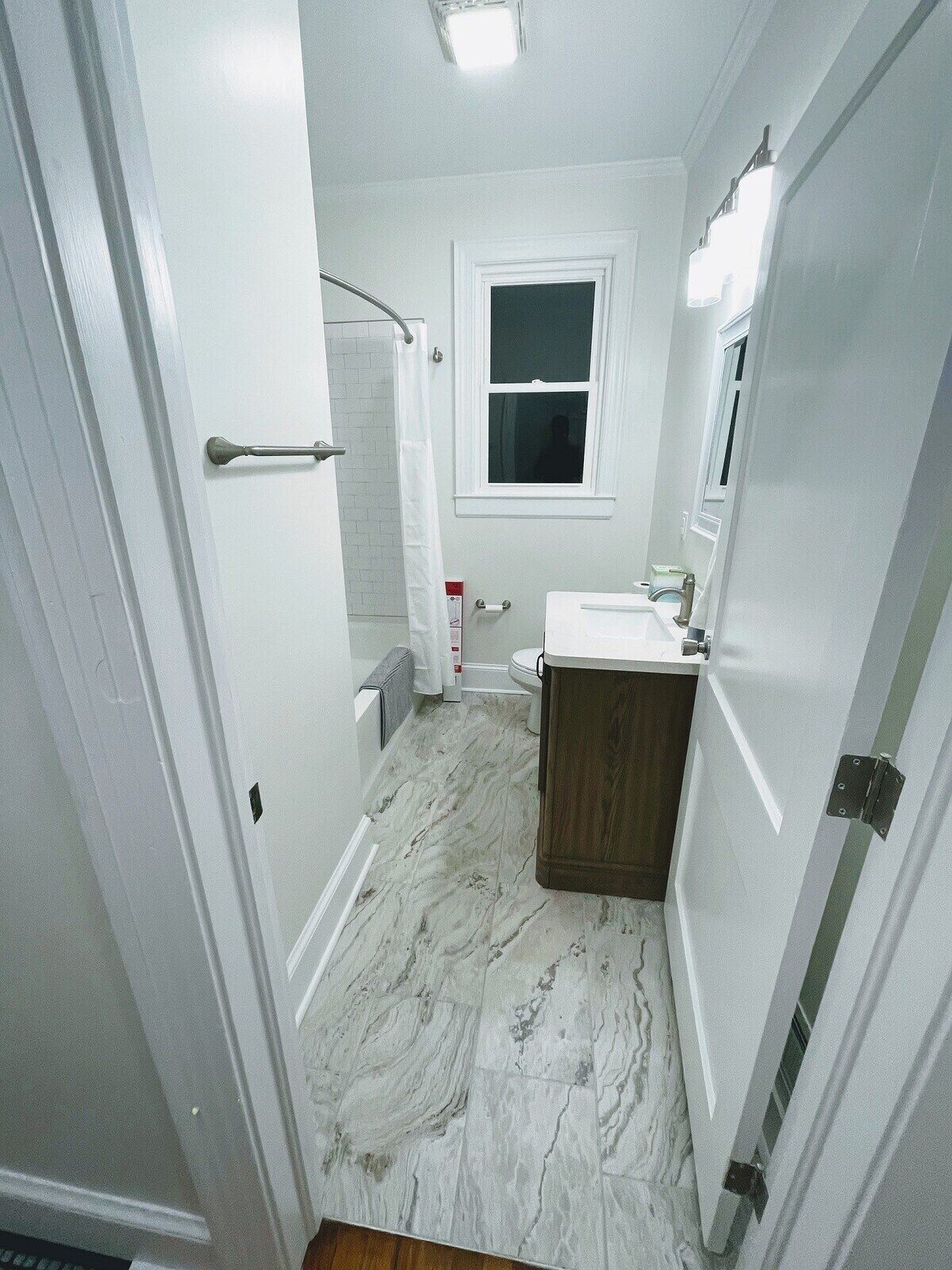 Un baño estrecho con paredes blancas, suelo de mármol, un inodoro blanco, una ventana pequeña y un armario de madera.