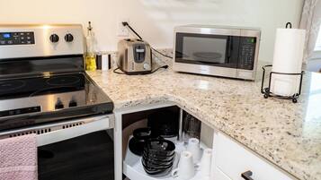 A compact kitchen with a stove, microwave, and a shelf above holding dishes and glasses.