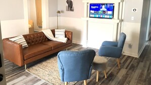 Een woonkamer met een bruine leren bank, twee blauwe fauteuils, een witte open haard en een televisie erboven.