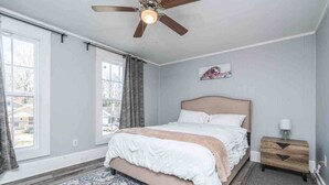 Een slaapkamer met een bed, een plafondventilator, een raam met gordijnen, een nachtkastje en een vloerkleed.
