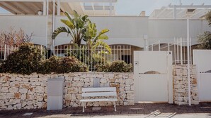 Eine weiße Bank vor einer Steinmauer mit einem Tor, einem weißen Zaun und einem Gebäude mit flachem Dach und einer Pergola.