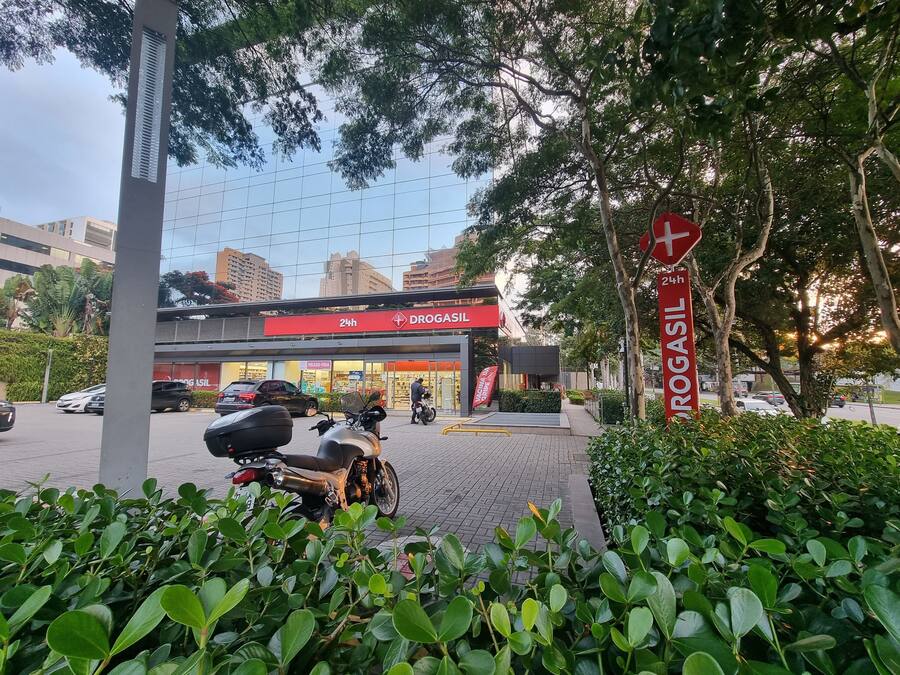 A 24-hour Drogasil store with a motorcycle parked in front, surrounded by greenery and a clear sky.
