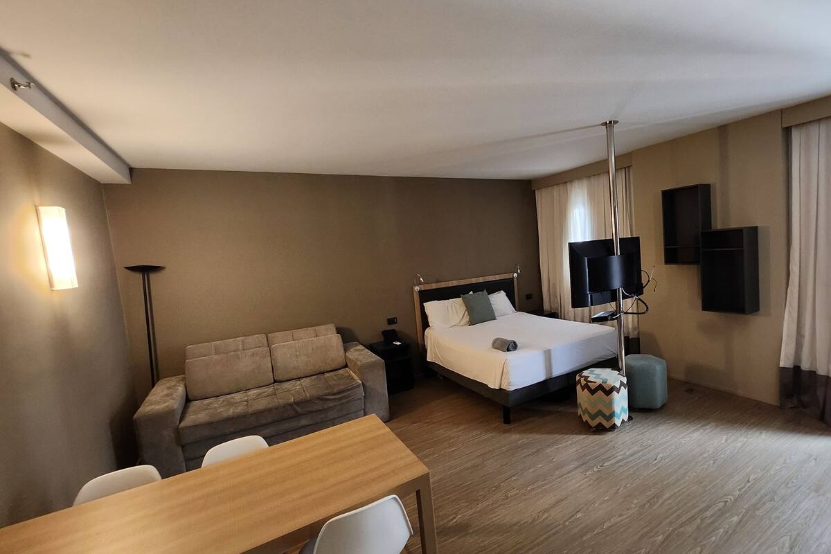 Une chambre d’hôtel avec un lit, un canapé, un bureau, une télévision et une fenêtre avec des rideaux.
